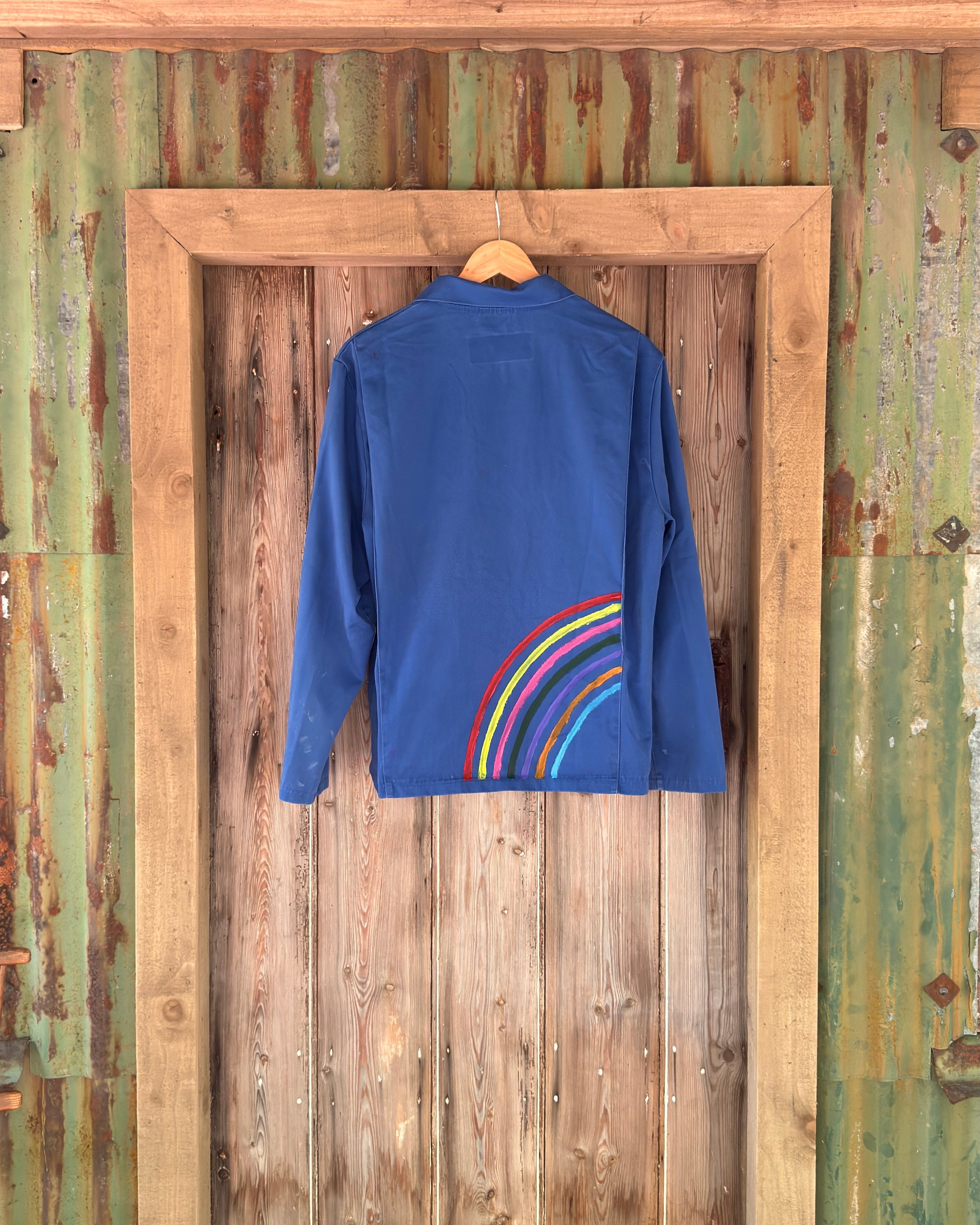 A royal blue long-sleeve shirt hanging on a wooden hanger against a rustic green corrugated metal door with a wooden frame.