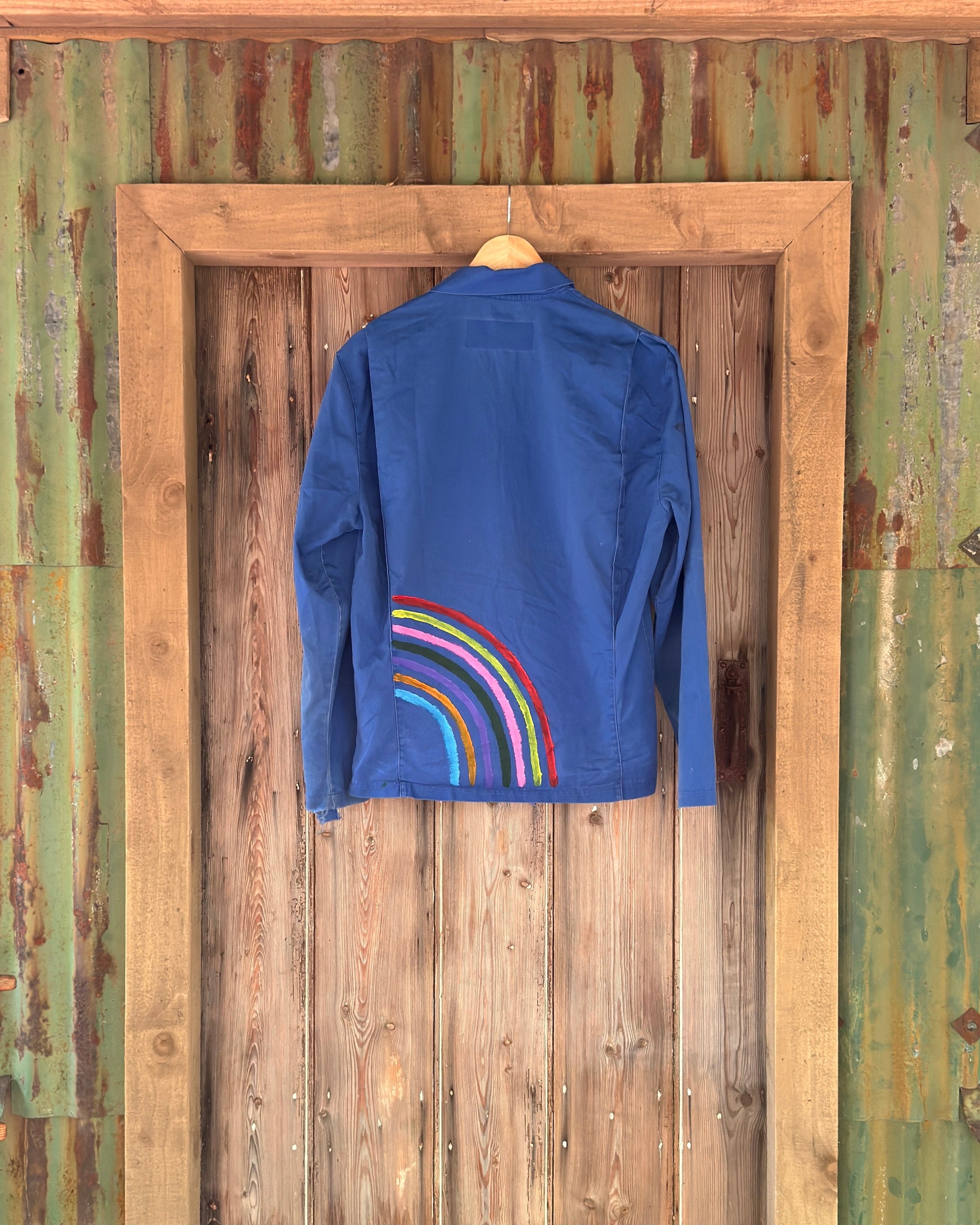 A blue long-sleeve shirt hanging on a wooden hanger against a rustic weathered green and brown wooden door, featuring a colorful rainbow graphic on the lower back.