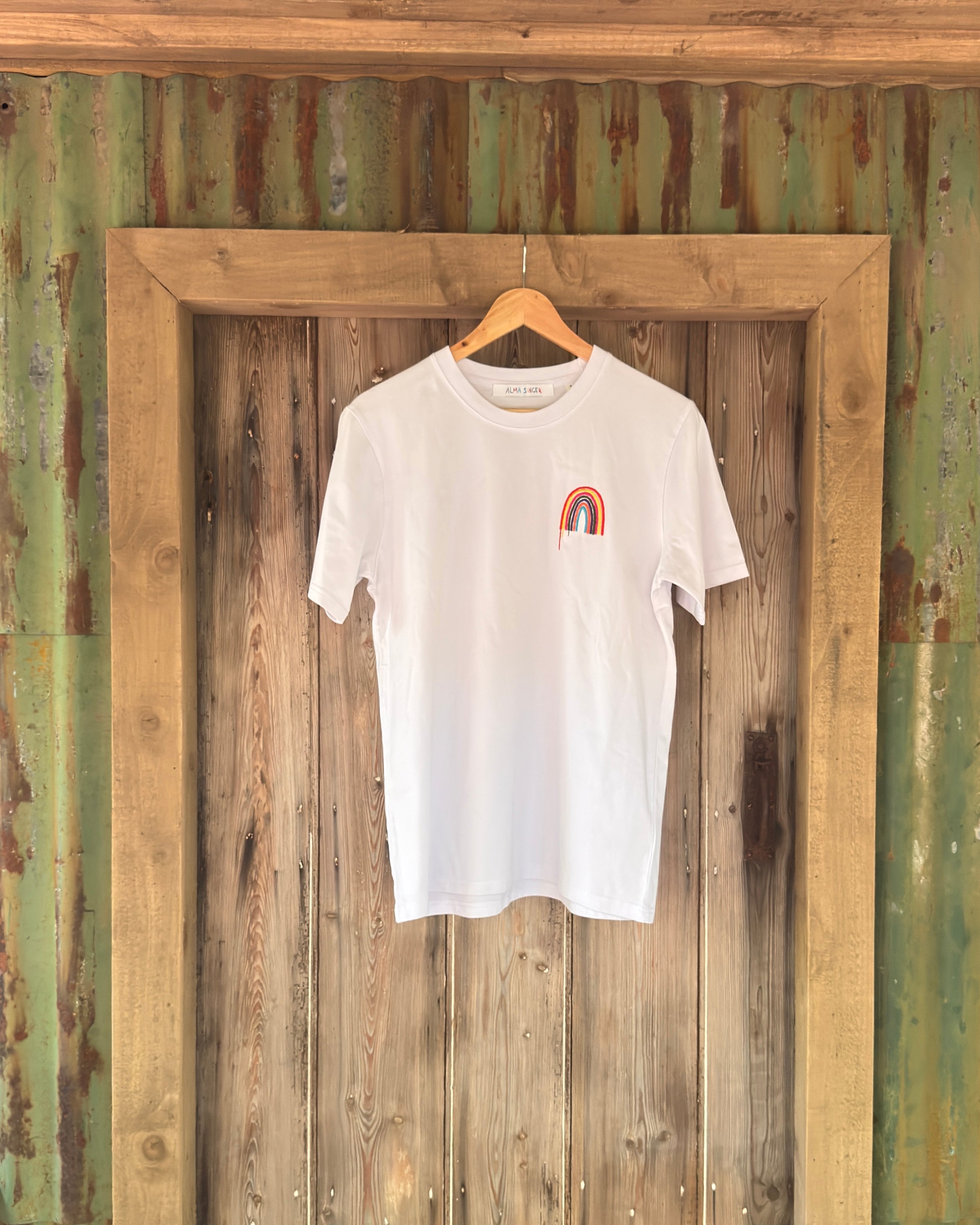 A white short-sleeve crewneck t-shirt with a small rainbow graphic on the left chest, hanging on a wooden hanger against a rustic wooden door and corrugated metal wall.
